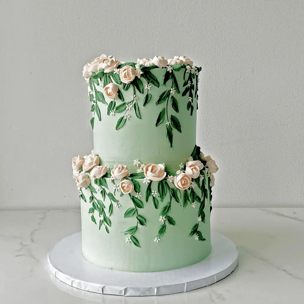 A two-tiered, mint green cake decorated with intricate green leaves and delicate light pink roses piped in frosting.