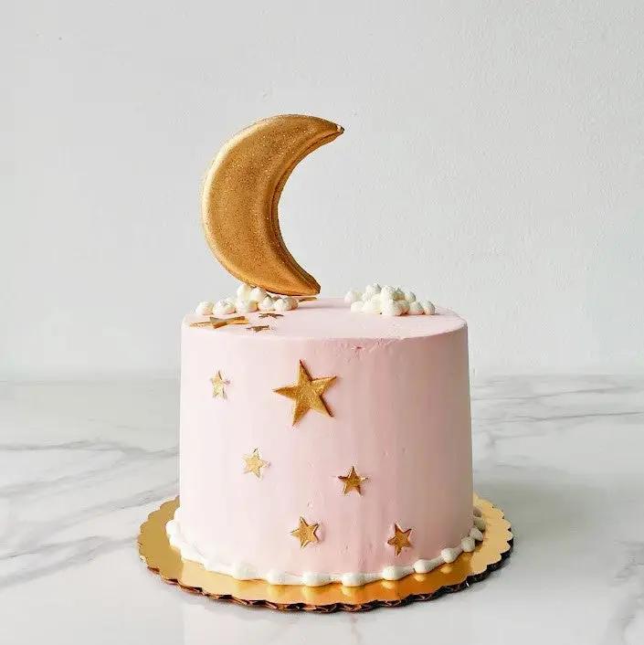 A pink cake with small clouds, gold stars and a crescent moon decoration at the top.