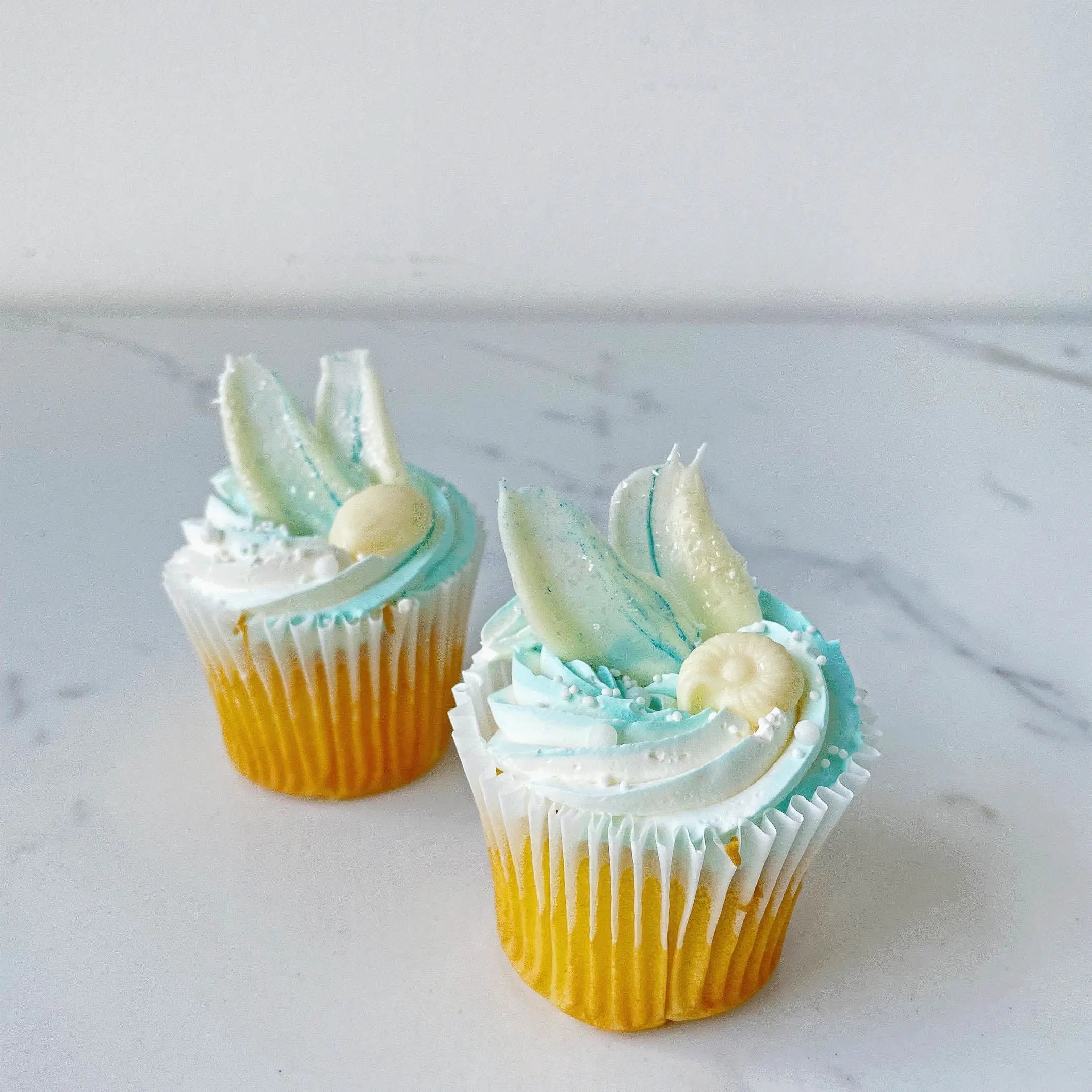 Two cupcakes with an orange base and white and light blue frosting, each topped with two white chocolate or fondant bunny ears.