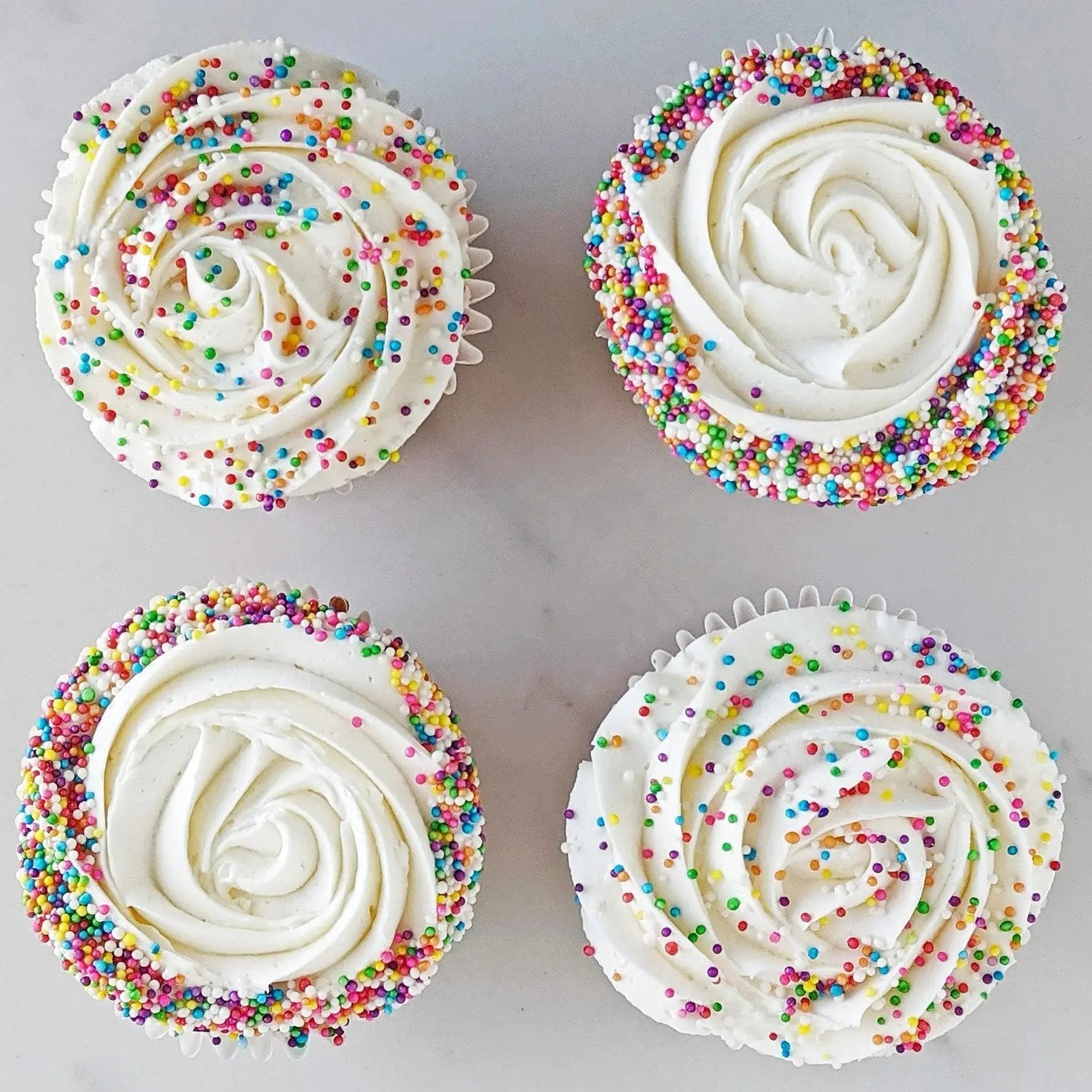 A photo of four cupcakes with white frosting and multicolored Rainbow sprinkles on top and around the base.