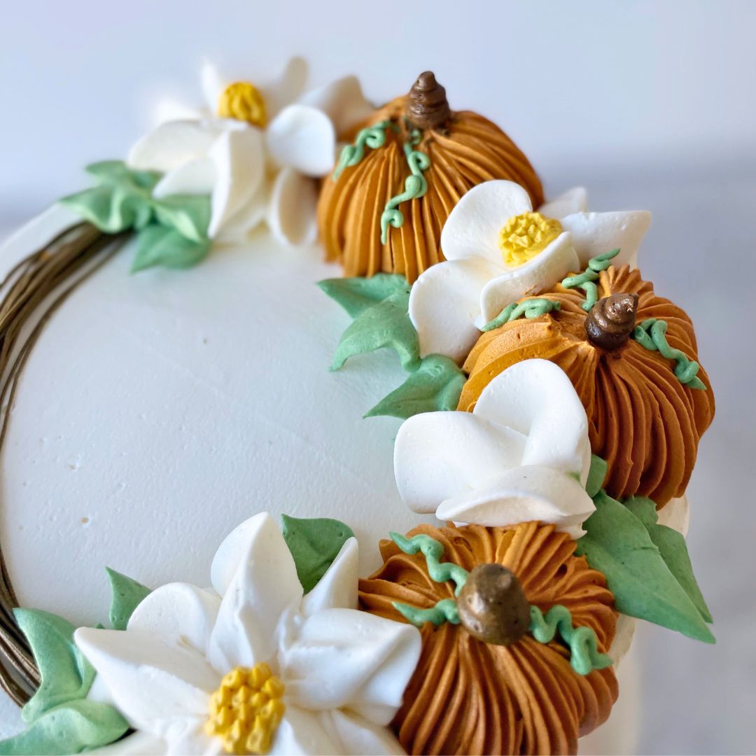 A white cake with a wreath of buttercream flowers and orange pumpkins on top.