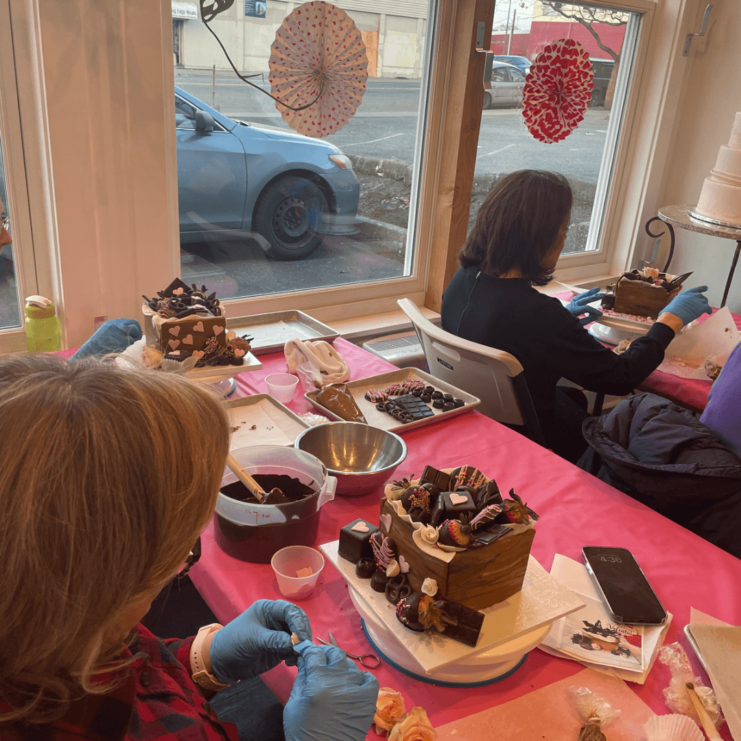 Two people in a baking class, each decorating a chocolate cake with chocolate-covered strawberries and other chocolate decorations.