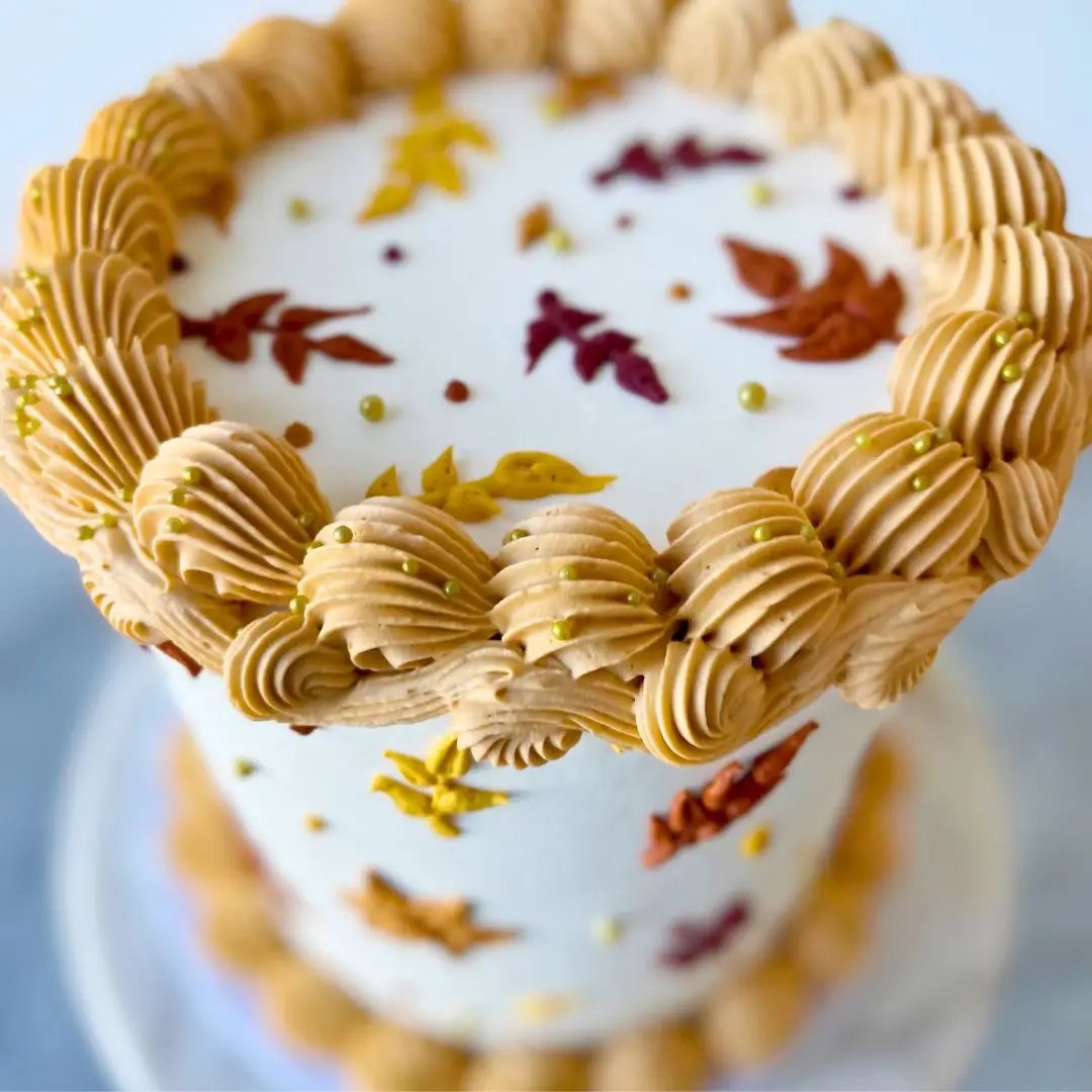 A small round cake with ridged piping on the top and bottom, and a design of colorful autumn leaves around the side.