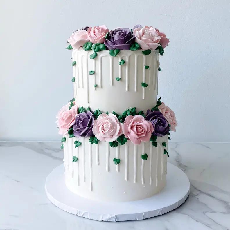 A two-tiered white cake. The sides of both tiers are decorated with vertical lines and clusters of pink and purple flowers at the top of every tier.