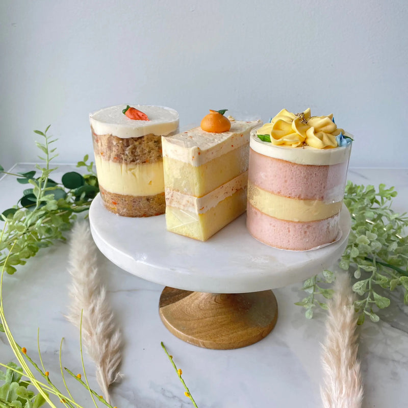 Three small, layered cakes displayed on a small white cake stand with the color of pink, yellow, brown, and white.