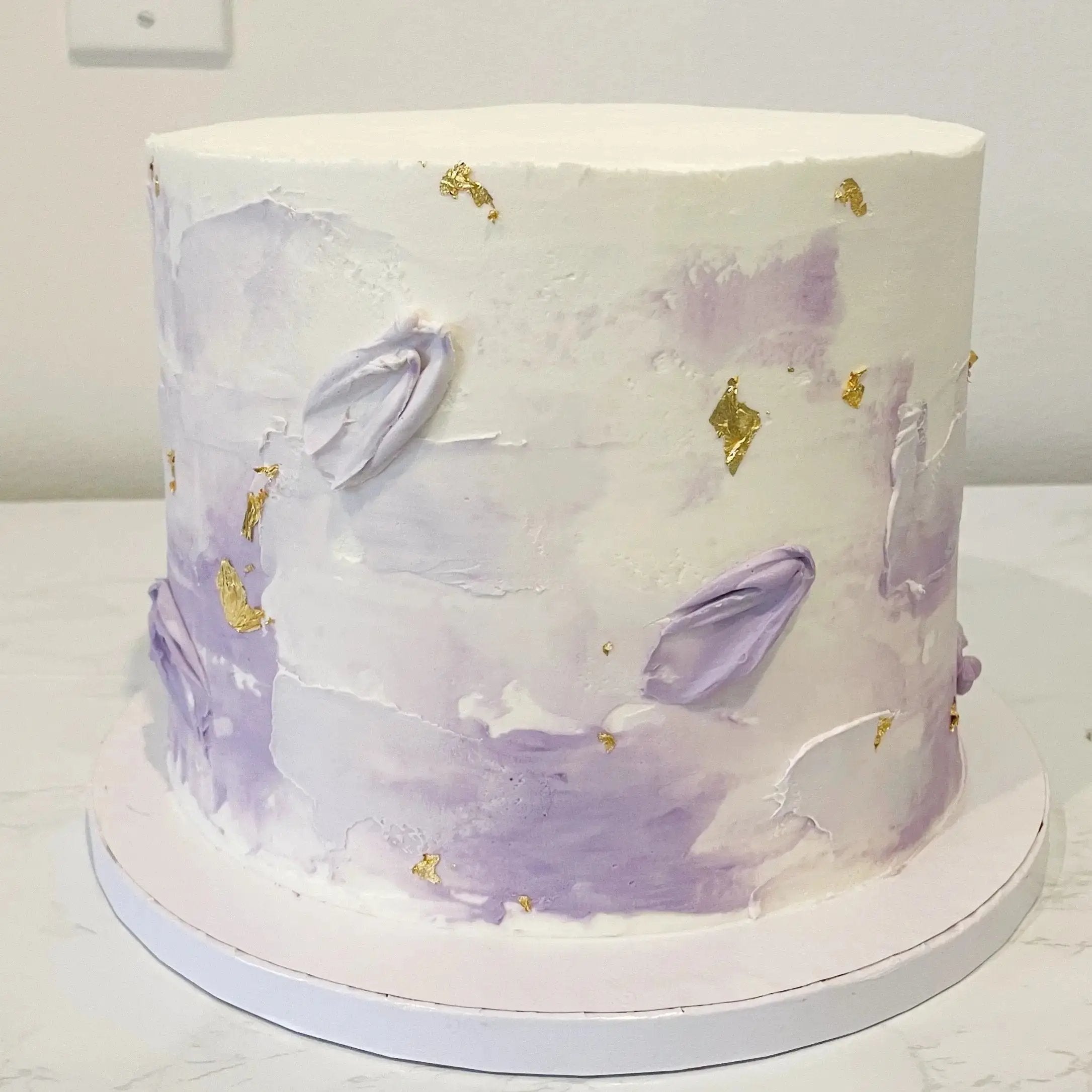 A short white cake with a textured, abstract design featuring light purple watercolor-like splotches, and flecks of gold leaf.