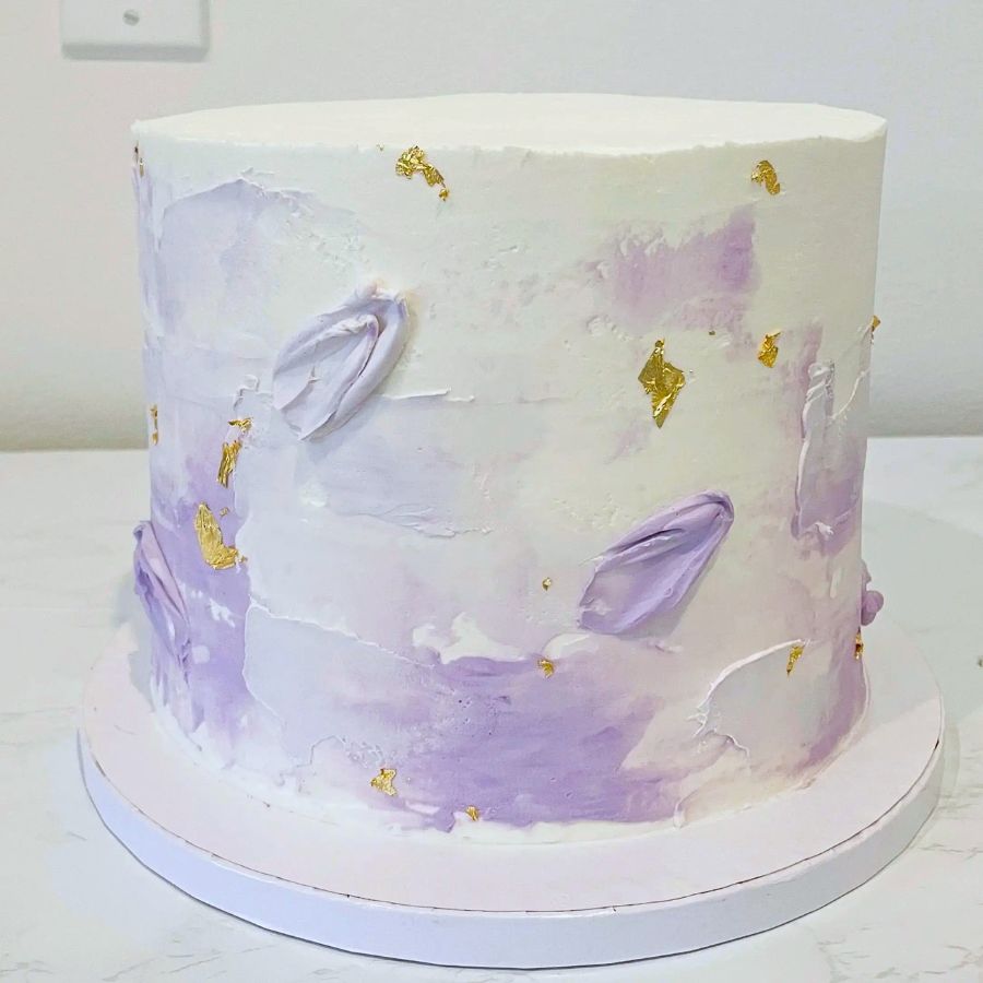 Cylindrical cake with purple and white floral decorations on a white surface.