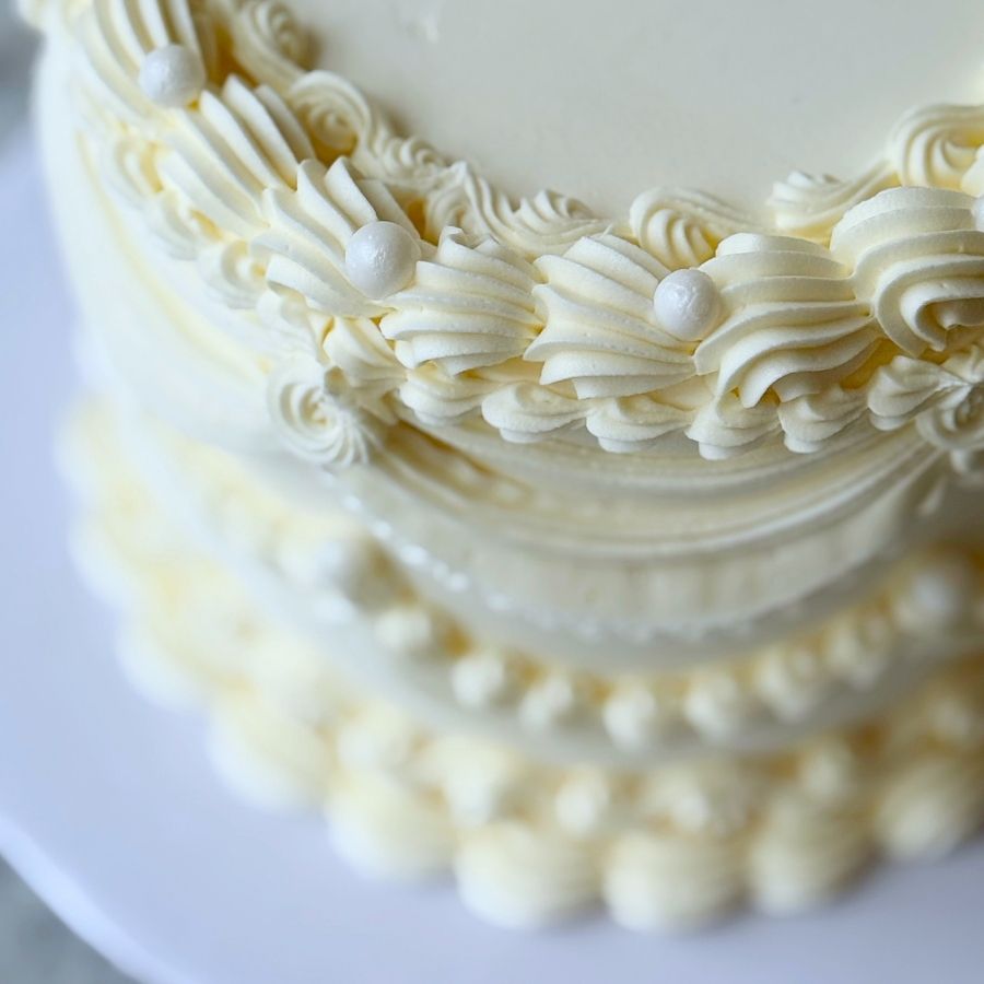 An overhead, close-up shot focusing on the top edge of a white frosted cake. The image highlights a thick, textured buttercream border with a shell-like pattern and decorative white pearls tucked into the folds of the icing.