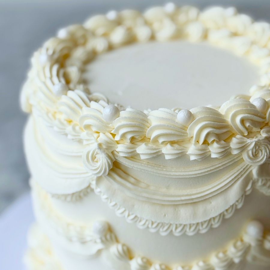 A soft-focus, close-up image of the upper rim of a white tiered cake. The detailed piping features swirling crests and round white sugar pearls, with the smooth top of the cake partially visible in the center.