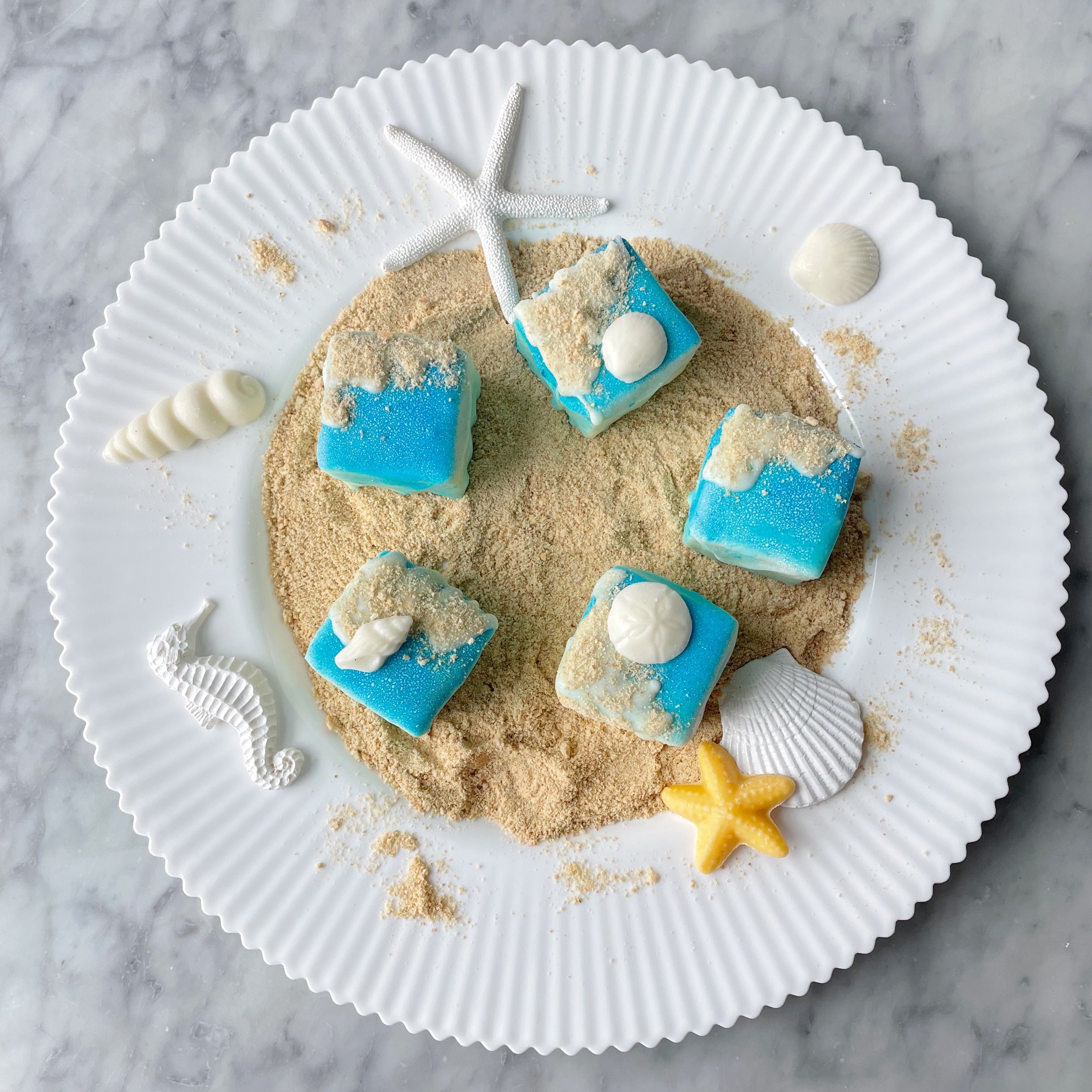A plate with white chocolate covered vanilla cakes designed to look like beach sand, complete with decorative seashells and a starfish.
