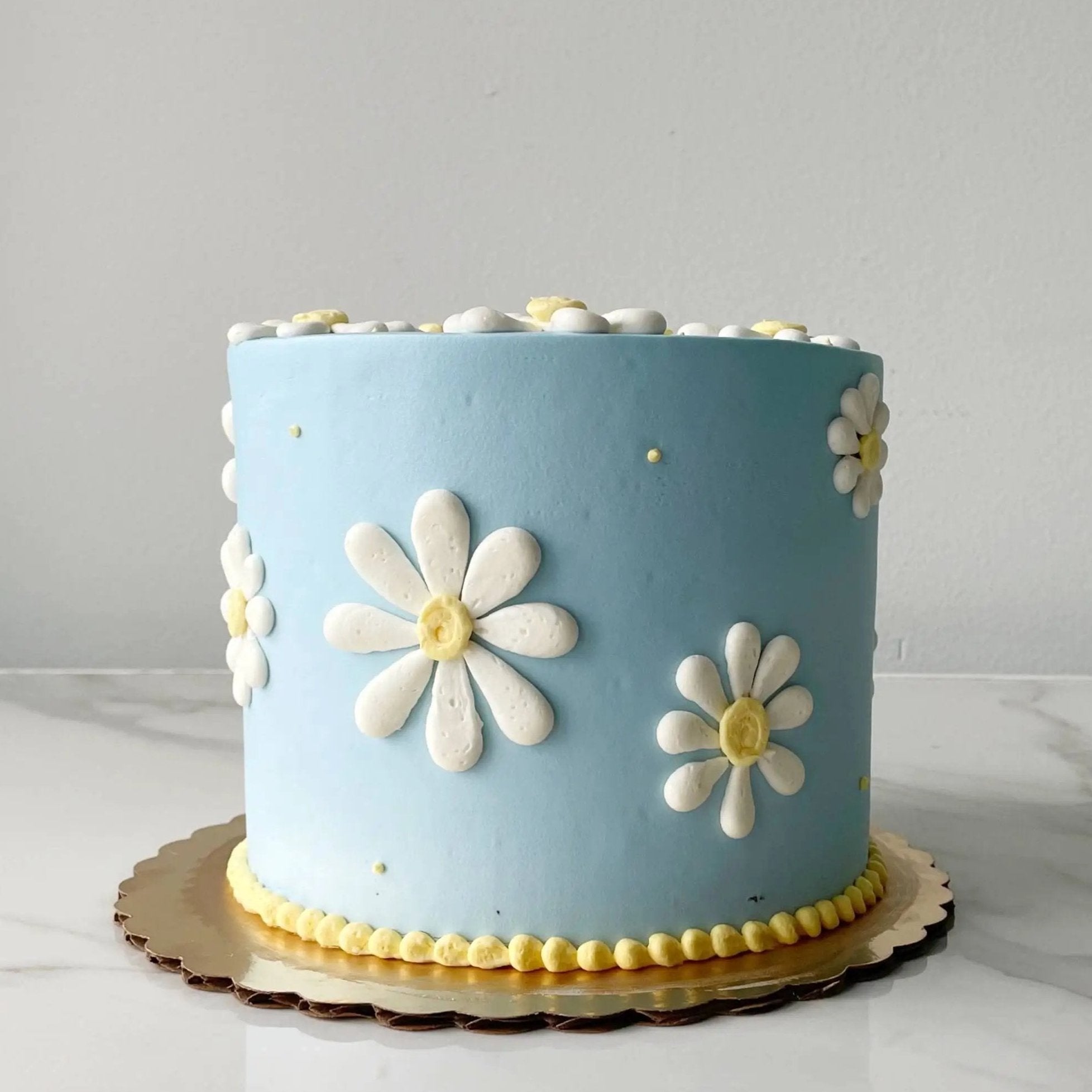 A smooth, light blue round cake. The side of the cake is decorated with white and yellow daisies of various sizes.