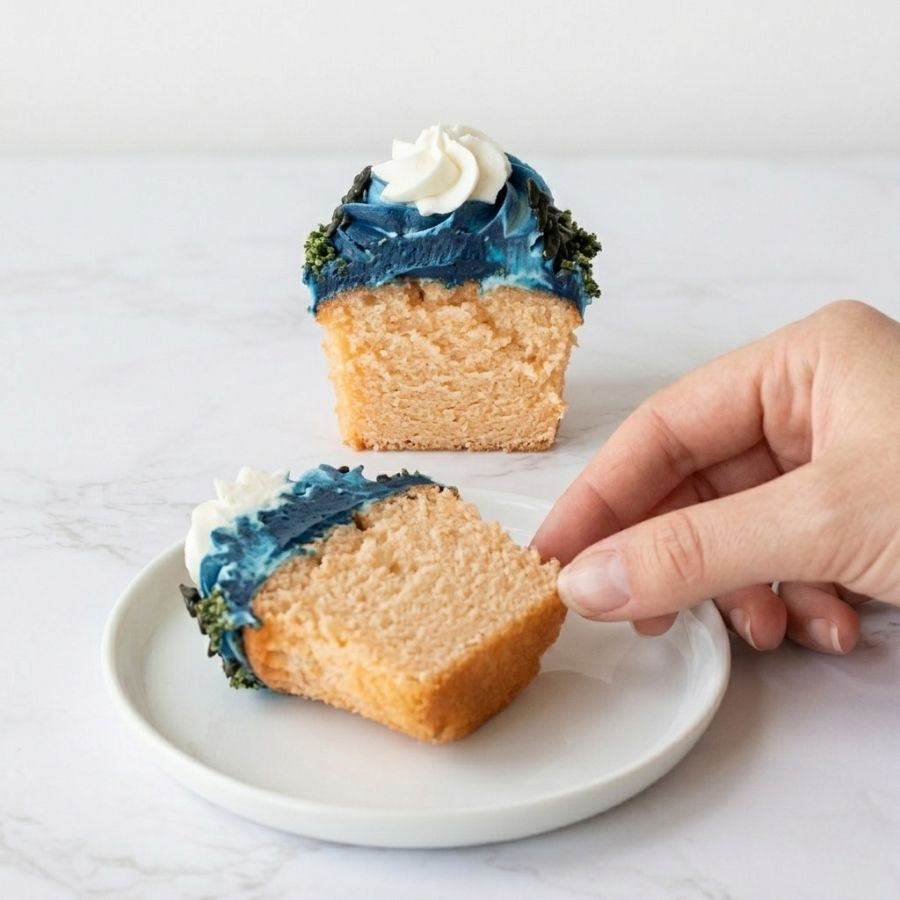 Half-eaten cupcake with blue and green frosting on a white plate, hand reaching for it.