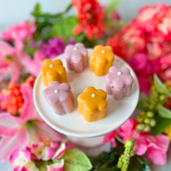 A selection of colorful daisy-shaped tea cakes on a white plate, displayed with a background of vibrant flowers.