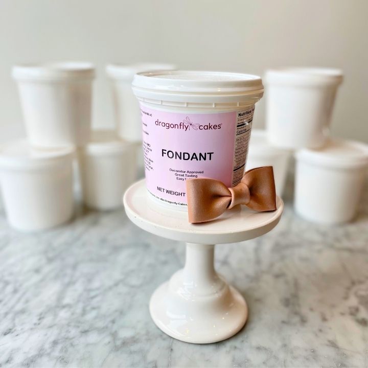 Container of dragonfly cakes fondant with a brown bow made from the fondant on a small white cake stand.