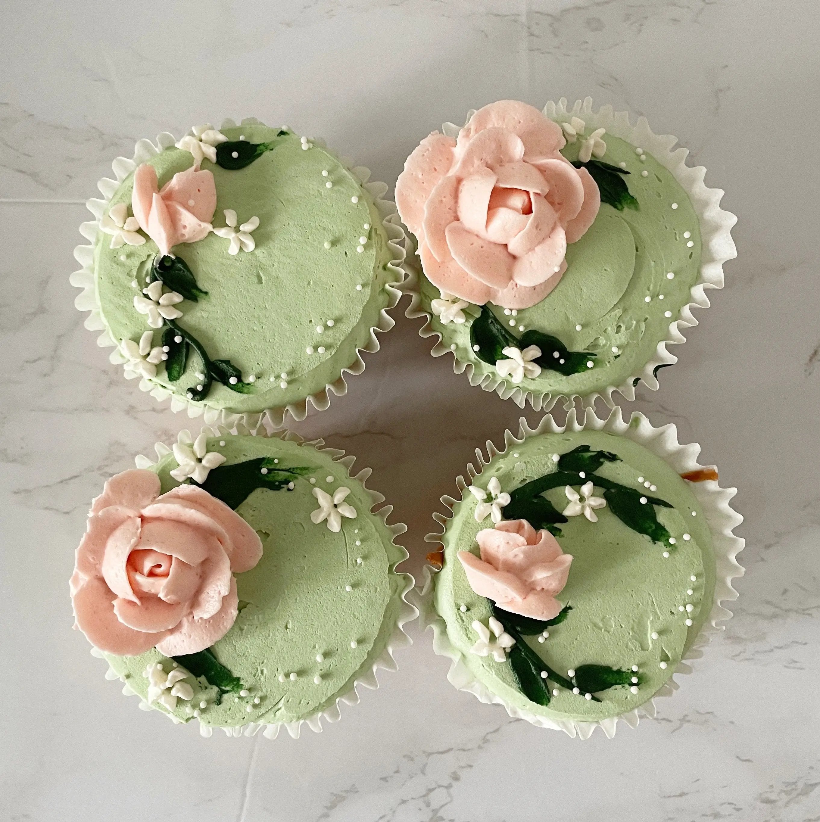 A top-down view of four cupcakes with light green frosting, each decorated with a pink rose and green leaves.