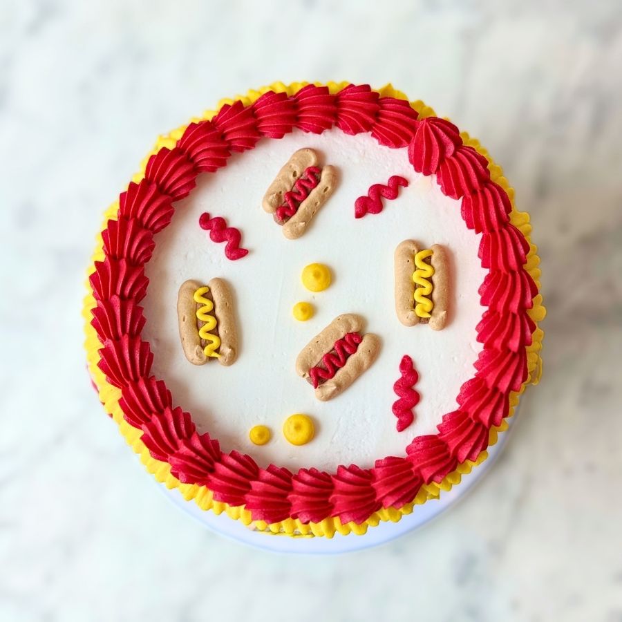 An overhead view of a round red cake. The top is smooth and surrounded by a thick border of red and yellow frosting ruffles. It is decorated with small white sugar pearls.
