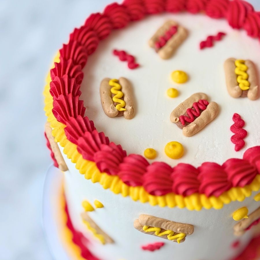 A close-up side view of a red cake. It is decorated with yellow frosting that looks like mustard and white frosting ruffles. There are small white and blue circle decorations on the side.