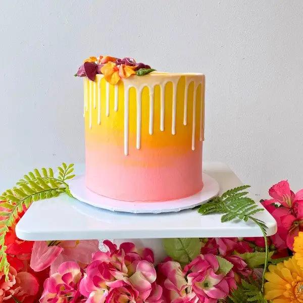 A Summer Sunset Drip Cake with orange-on-orange mousse, yellow-to-pink buttercream gradient, and white chocolate drip, garnished with seasonal blooms on top, presented on a white plate with floral background.