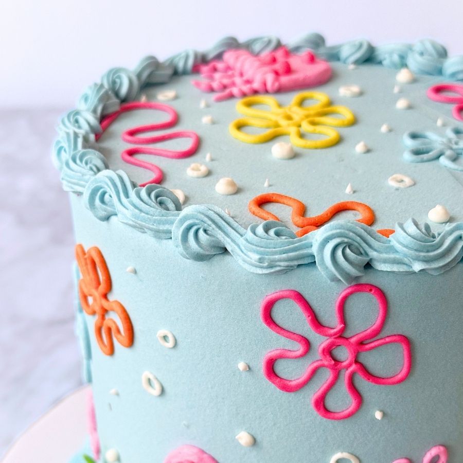 A close-up, angled view of the top edge of a light blue cake. The top is decorated with piped buttercream flower shapes in pink, orange, and yellow, along with small white bubbles. A textured blue wave-like border runs along the upper rim.