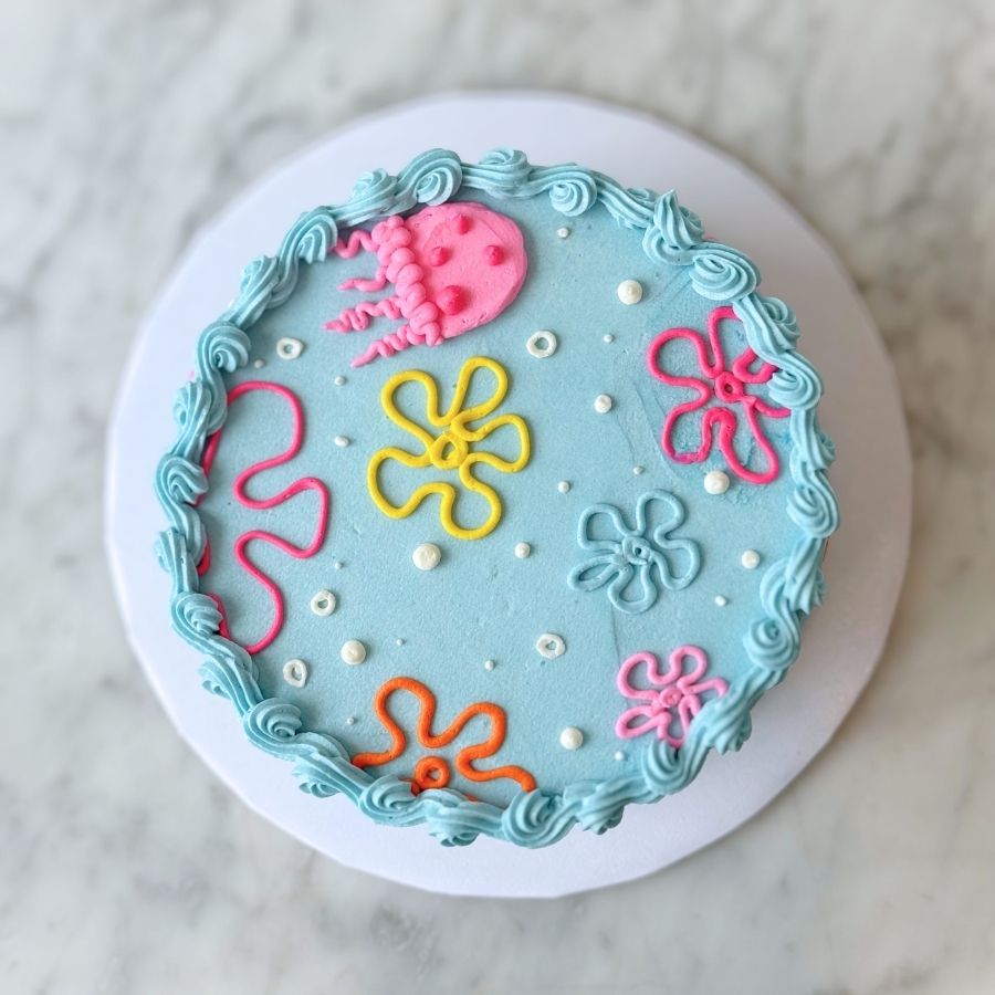 An overhead view of a round cake with a smooth light blue surface. The top is covered in whimsical buttercream designs, including a pink jellyfish and several multi-colored flower outlines in yellow, pink, and orange, all framed by a blue scalloped border.