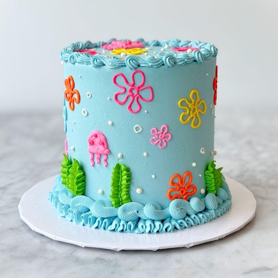 A full-view shot of a tall, light blue cake themed after an underwater world. The cake is decorated with vibrant pink jellyfish, green seaweed ruffles at the bottom, and colorful floral patterns. It sits on a white cake board against a clean, bright background.