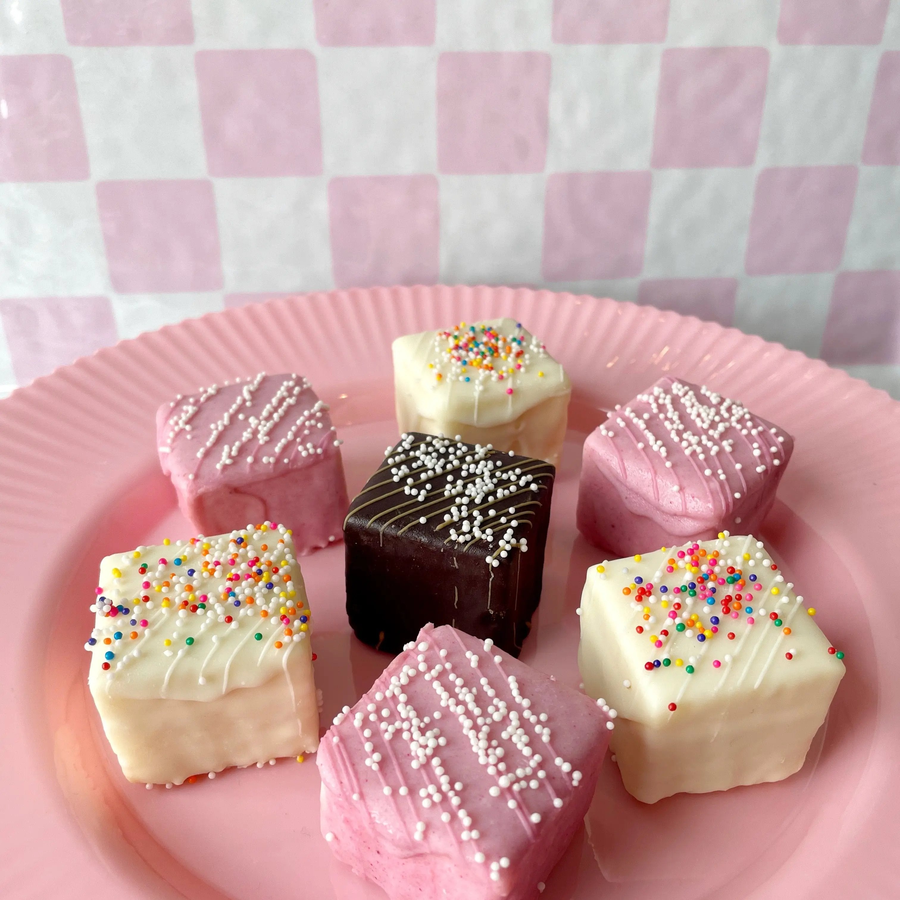 Assorted petit fours with sprinkles on a pink plate against a pink checkered background