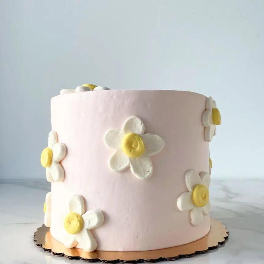 A smooth, light pink round cake. The side of the cake is decorated with white and yellow daisies of various sizes.