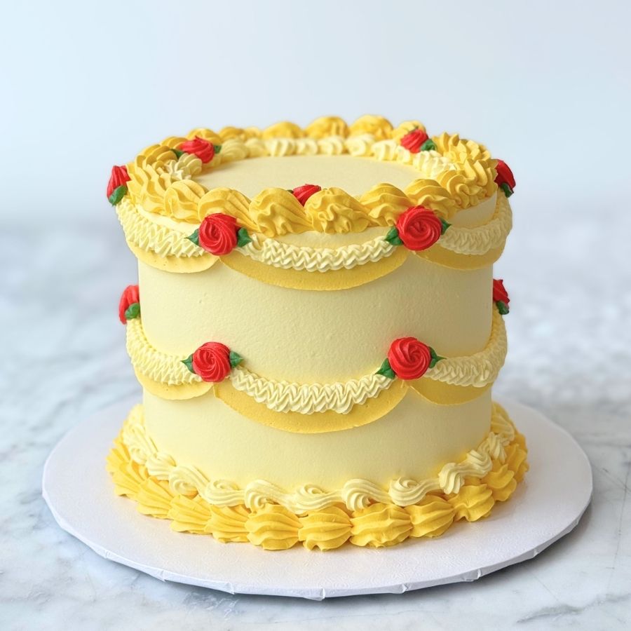 A full-view shot of a tall, bright yellow cake inspired by a classic fairy tale. The cake is heavily decorated with gold-toned buttercream swags, red rose accents, and gold pearls. It is displayed on a white cake board over a marble surface against a neutral background.