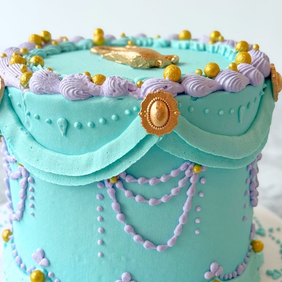 A close-up side view of the "Princess Genie" cake, highlighting the intricate gold-dusted buttercream ruffles and teal scale-like piping. Several small pink sugar flowers and shimmering gold pearls are nestled within the ornate icing details.