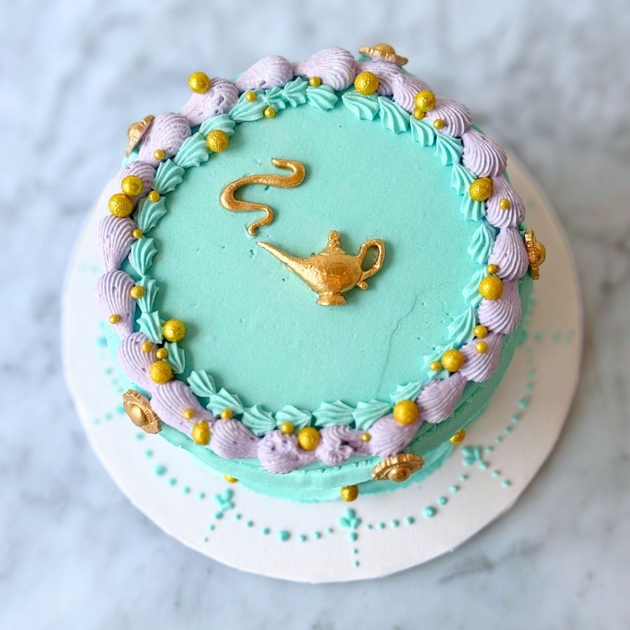An overhead view of a round teal cake with a smooth top. The outer rim is decorated with a dense, regal crown of gold and teal buttercream swirls, embellished with pink flower accents and gold sugar beads.
