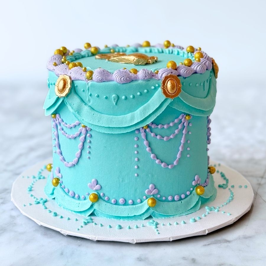 A full view of a tall, vibrant teal cake inspired by a desert princess. The cake features elaborate gold and teal buttercream piping, including horizontal swags and vertical ruffles. Gold sugar pearls and small pink blossoms accent the design, set on a white board against a marble background.