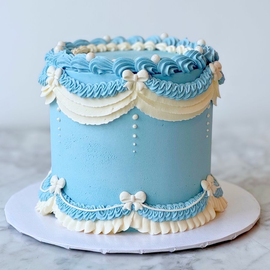 A full view of a tall light blue cake on a white stand. The cake has fancy white and blue frosting designs, gold beads, and pink flowers. It is shown against a plain white background on a marble table.