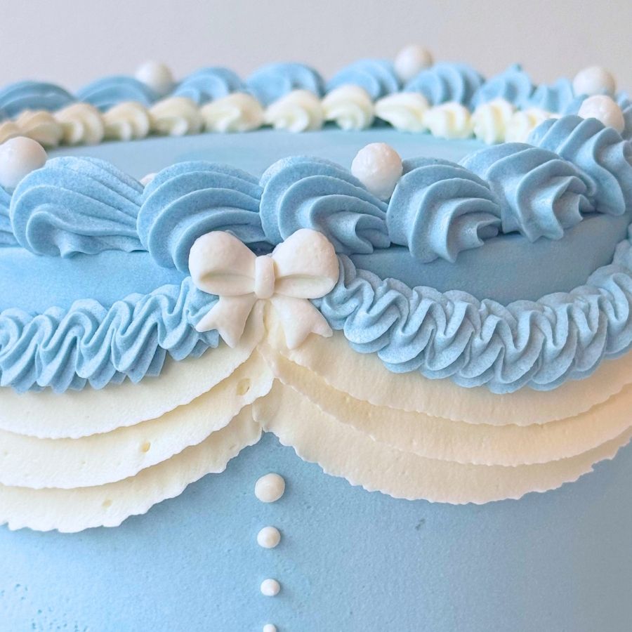 A close-up side view of a light blue cake. It is decorated with white frosting ruffles, gold beads, and small pink flowers. A large white frosting bow with a gold center is on the side.