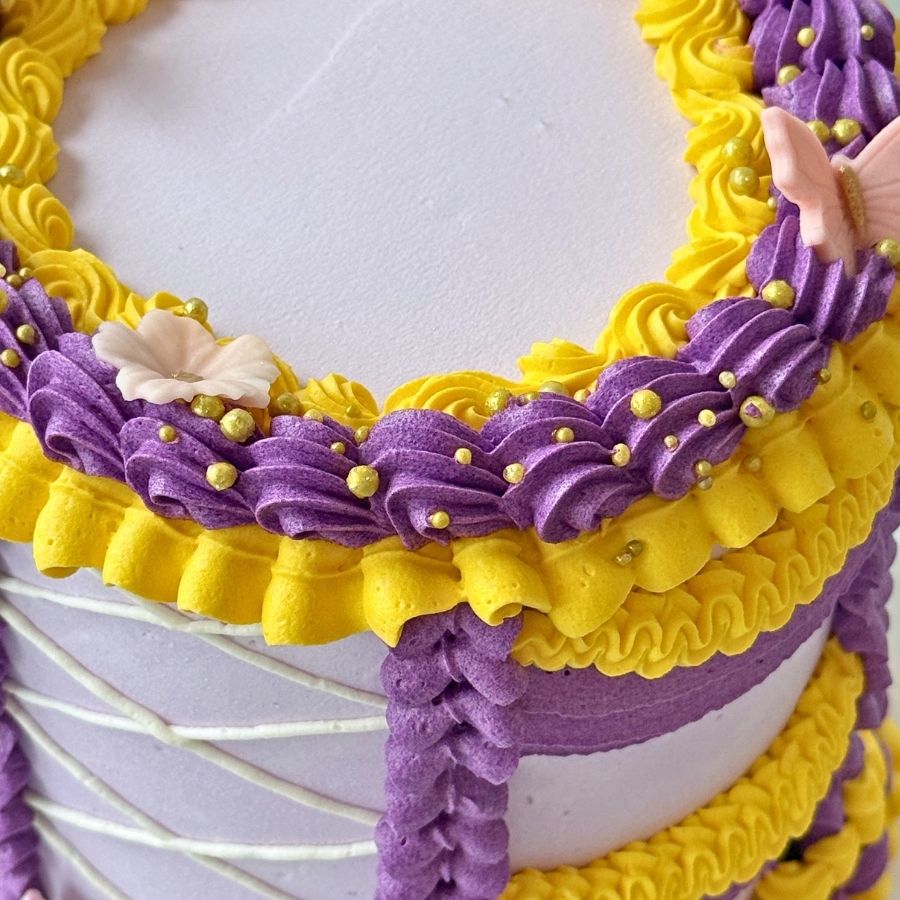 An overhead view of a round cake with smooth lavender frosting on top. The outer edge is decorated with a thick, ornate crown of purple and yellow buttercream ruffles, featuring gold pearls, small pink blossoms, and sugar butterflies.
