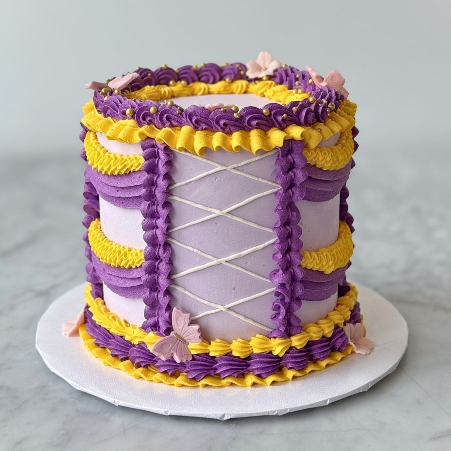 A full-view shot of a tall, whimsical princess-themed cake. The lavender cake is decorated with purple and yellow buttercream swags, a white piped corset detail on the front, and pink butterfly and flower accents. The cake sits on a white board over a marble surface.