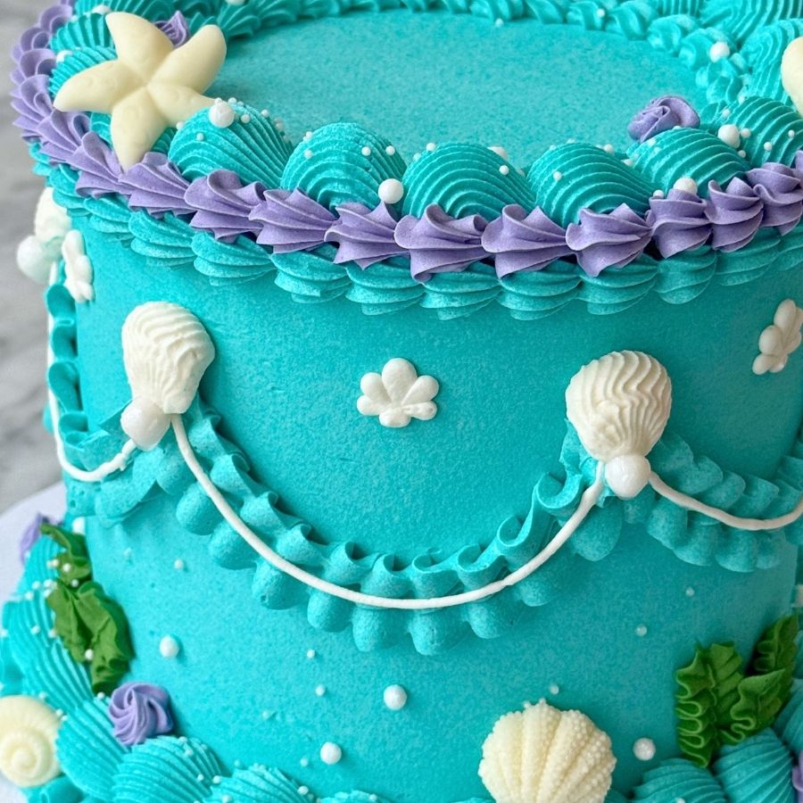 A close-up side view of a teal and purple ombre cake. The design features intricate piping that resembles scales and sea waves, decorated with edible gold pearls, small pink sugar flowers, and a large white seashell accent.