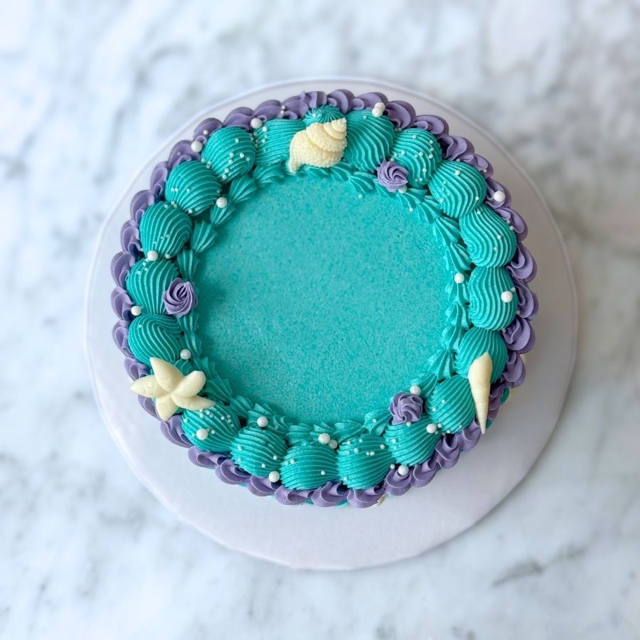 A macro detail shot of the side of a mermaid cake. It showcases vertical purple buttercream ruffles alongside teal scale-like piping. The texture is accented with shimmering gold pearls and a small, realistic white sugar starfish.