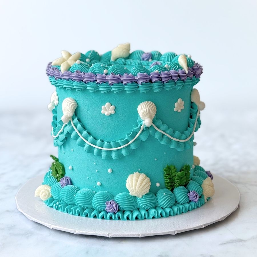 A full-length view of a tall, elegantly decorated mermaid princess cake. The cake transitions from deep purple at the base to soft teal at the top, featuring elaborate wave piping, gold accents, and assorted sea-themed sugar art. It is presented on a white cake board over a marble tabletop.