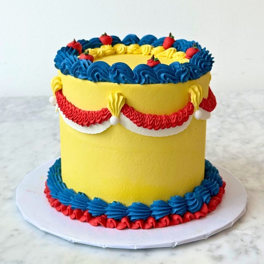 A full view of a tall dark blue cake on a white board. The cake has red and blue frosting designs, gold beads, and a red apple decoration on the front. It sits on a marble table.