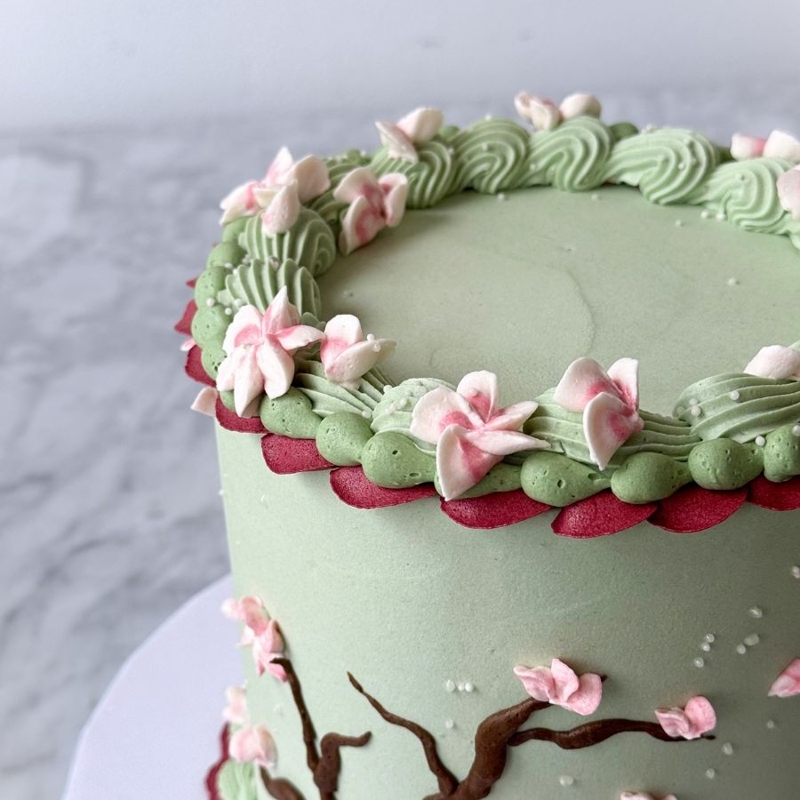 A perspective shot focusing on the top rim and side of a green cake. It showcases the detail of the pink buttercream flowers tucked into sage green frosting ruffles, positioned above a contrasting dark red scalloped trim. The cherry blossom branch design is visible on the side.