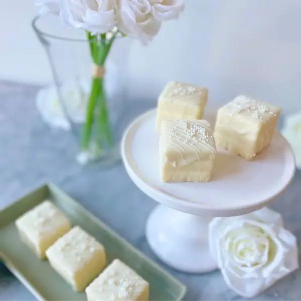 white tea cakes with creamy white chocolate coating and edible pearl dragees on a white platter, with a vase of white flowers and a larger cake in the background.