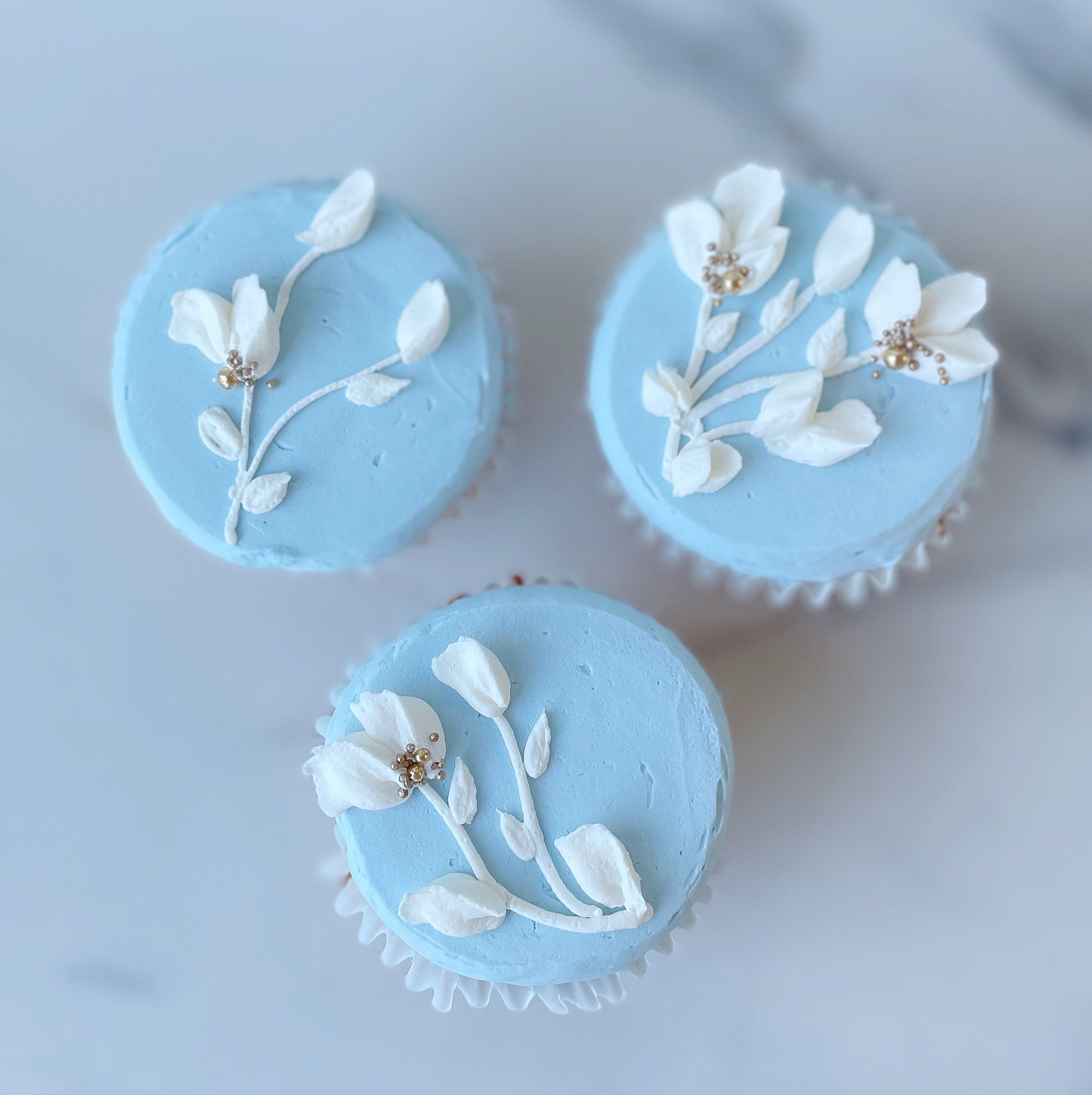A set of three cupcakes with blue icing and white flower decorations, placed on a marble surface.