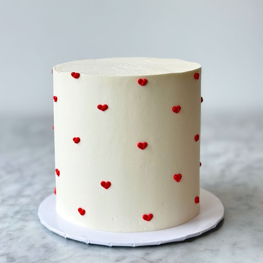 A tall, minimalist round cake covered in smooth off-white buttercream frosting. The sides are decorated with a delicate pattern of tiny, hand-piped red hearts. The cake is centered on a white cake board and set against a neutral, bright background on a marble surface.